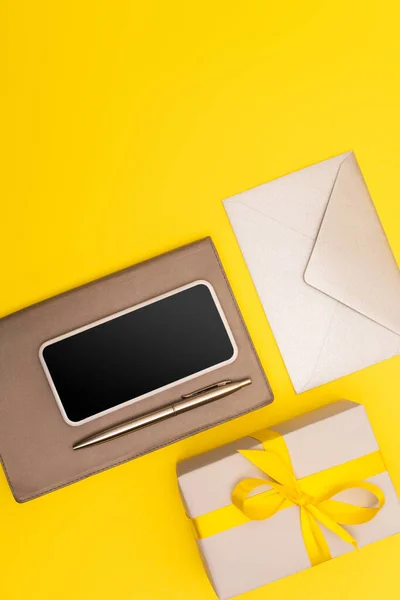 Top view of smartphone with blank screen on copy book with pen near envelope and present isolated on yellow — Stock Photo