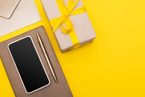 Vista superior do smartphone com tela em branco no livro de cópia com caneta perto do envelope e caixa de presente isolada no amarelo — Fotografia de Stock