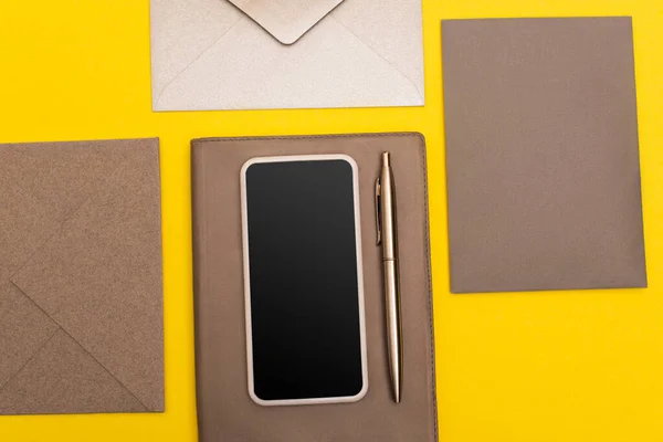 Vista dall'alto dello smartphone con schermo bianco su libretto di copie con penna vicino buste isolate su giallo — Foto stock