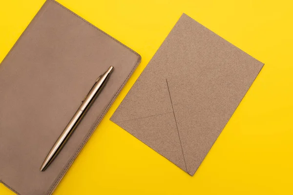 Верхний вид ноутбука с золотой ручкой рядом с бумажным конвертом изолирован на желтый — стоковое фото