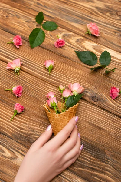 Cropped view of woman holding waffle cone with pink tea roses on wooden surface — Stock Photo