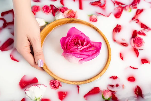 Vue recadrée d'une femme tenant un bol en bois avec pivoine rose près des roses dans de l'eau laiteuse — Photo de stock