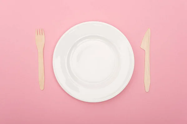 Top view of disposable wooden cutlery near white plate isolated on pink — Stock Photo