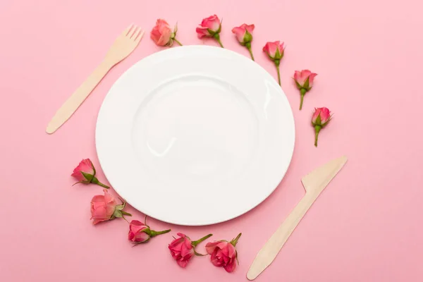 Vista de ángulo alto de cubiertos de madera desechables cerca de plato blanco y flores aisladas en rosa - foto de stock