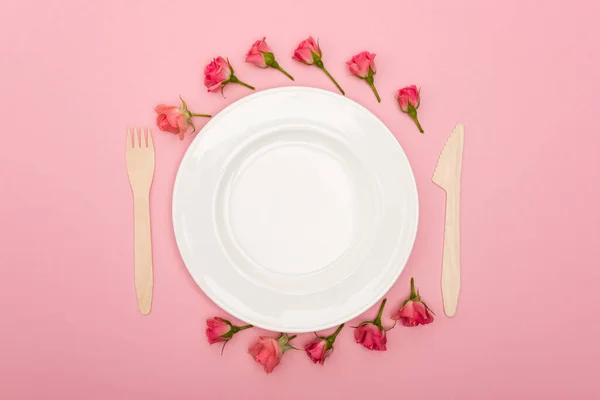 Flat lay with disposable wooden cutlery near white plate and tea roses isolated on pink — Stock Photo