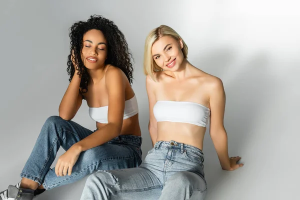 Улыбающиеся мультикультурные женщины в джинсах и белых топах сидят на сером фоне — стоковое фото