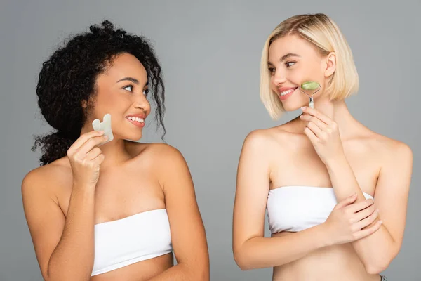 Smiling multiethnic women in white tops using jade roller and gua sha isolated on grey — Stock Photo