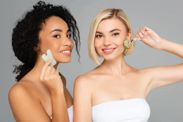 Mujeres multiétnicas con la piel perfecta masajeando caras con gua sha y rodillo de jade aislado en gris — Stock Photo