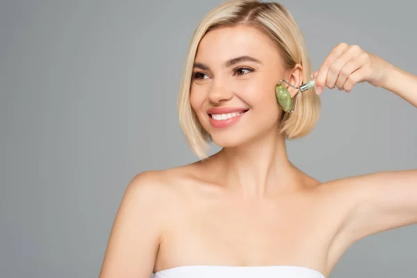 Donna sorridente con pelle perfetta massaggio viso con rullo di giada isolato su grigio — Foto stock