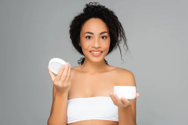 Sorrindo mulher americana africana em frasco branco top segurando com creme cosmético isolado em cinza — Fotografia de Stock