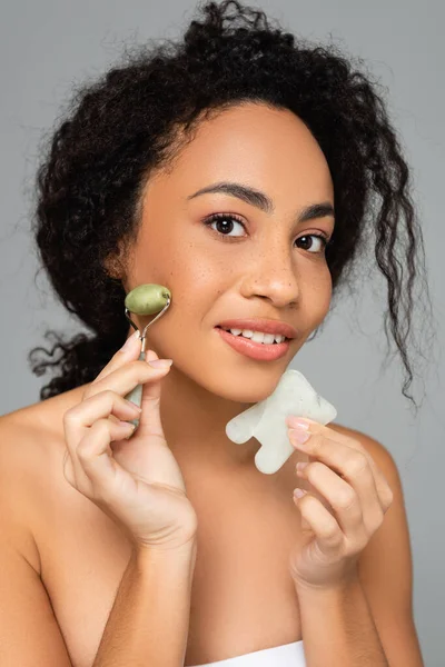 Mujer afroamericana sosteniendo rodillo de jade y gua sha cerca de la cara aislada en gris - foto de stock