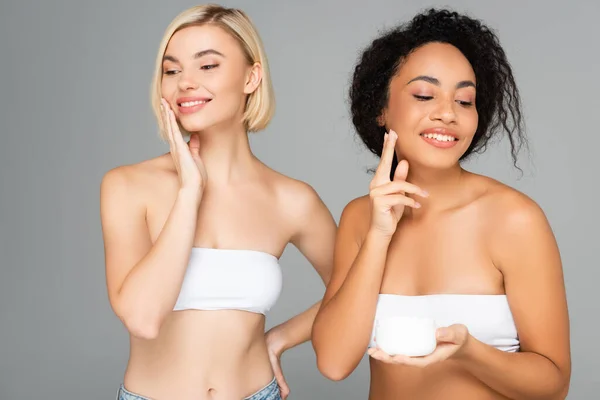 Femmes multiethniques gaies en hauts blancs appliquant de la crème cosmétique sur les visages isolés sur le gris — Photo de stock