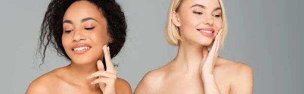 Sonrientes mujeres multiétnicas con hombros desnudos aplicando crema cosmética aislada en gris, pancarta - foto de stock