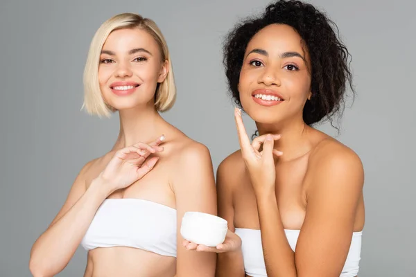 Smiling interracial women with perfect skin holding cosmetic cream isolated on grey — Stock Photo
