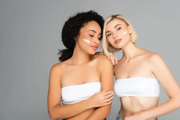 Blonde woman standing near african american woman with cream on cheek isolated on grey — Stock Photo