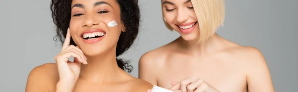 Fröhliche multikulturelle Frauen, die kosmetische Creme isoliert auf grau auftragen, Banner — Stockfoto