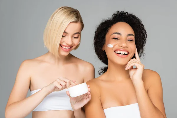 Веселі мультикультурні жінки з чистою шкірою нанесення косметичного крему на обличчя ізольовані на сірому — стокове фото