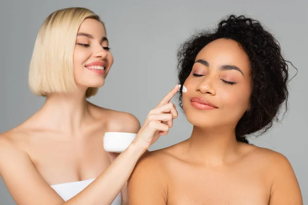 Femme afro-américaine debout près de femme blonde appliquant la crème cosmétique sur fond flou isolé sur gris — Photo de stock