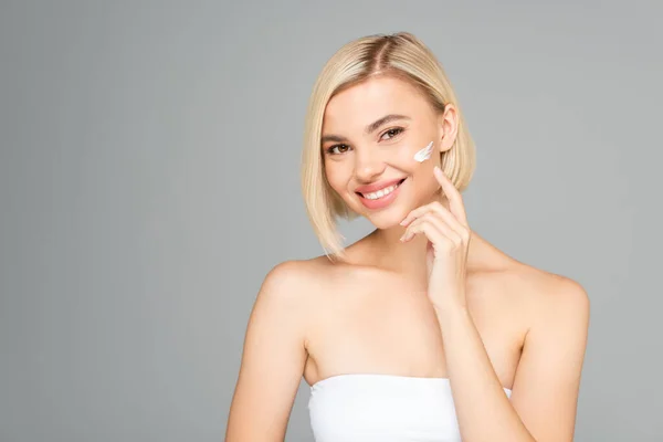 Mujer alegre sonriendo a la cámara mientras aplica crema cosmética aislada en gris — Stock Photo