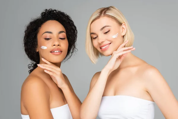 Interracial women with cosmetic cream and hands near faces isolated on grey — Stock Photo