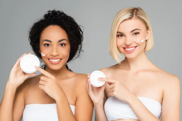 Cheerful multiethnic women holding jars with cream isolated on grey — Stock Photo
