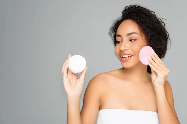 Young african american woman holding silicone brush and looking at cosmetic cream isolated on grey — Stock Photo