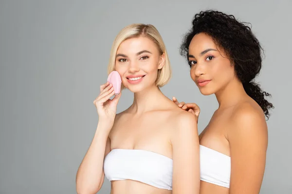 Jeunes femmes multiethniques en hauts blancs posant avec un nettoyant silicone isolé sur gris — Photo de stock