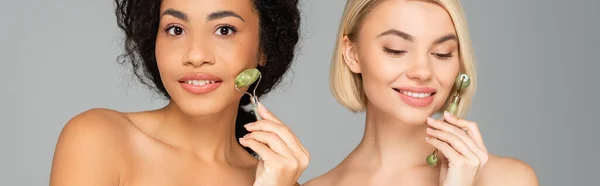 Young multicultural women using jade rollers isolated on grey, banner — Stock Photo