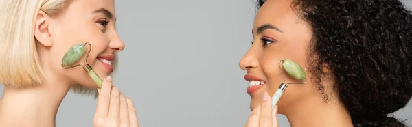 Side view of interracial women holding jade rollers near cheeks isolated on grey, banner — Stock Photo