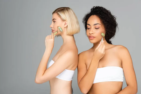 Young interracial women in white tops massaging faces with jade rollers isolated on grey — Stock Photo