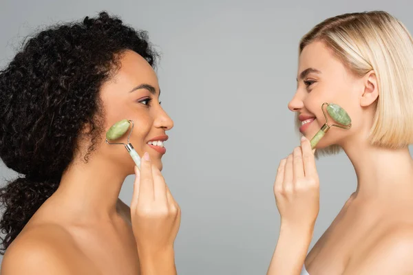 Side view of multiethnic women smiling at each other while using jade rollers isolated on grey — Stock Photo