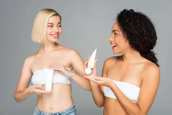 Smiling multiethnic women holding tubes of cosmetic lotion isolated on grey — Stock Photo