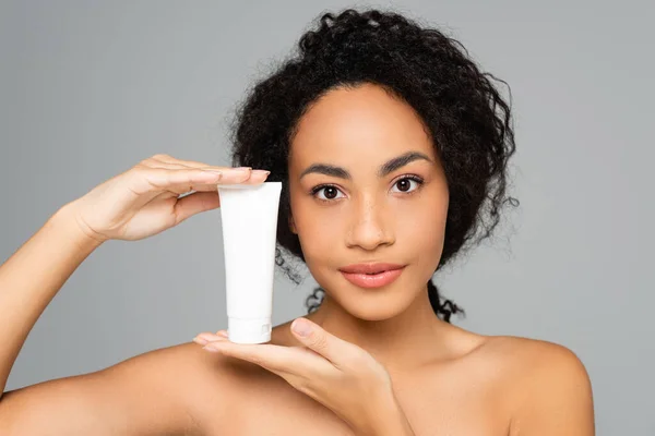 Joven mujer afroamericana con hombros desnudos sosteniendo tubo con crema cosmética aislada en gris - foto de stock