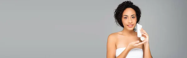 Femme afro-américaine souriante tout en tenant tube avec lotion cosmétique isolée sur gris, bannière — Photo de stock