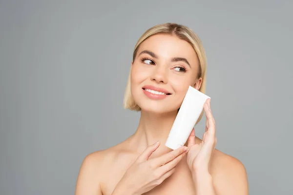 Mulher sorridente com ombros nus segurando tubo com creme cosmético perto do rosto isolado em cinza — Fotografia de Stock