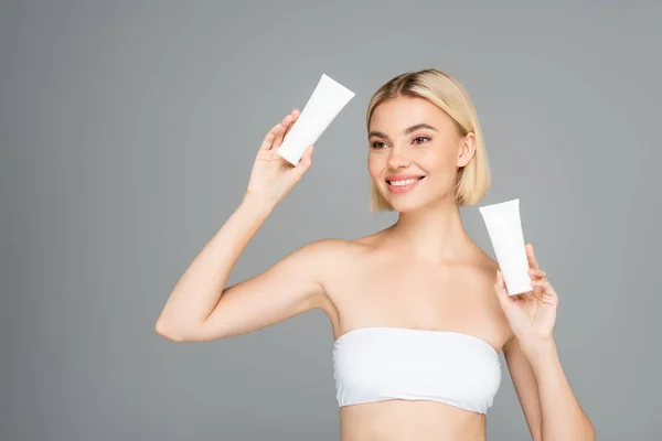 Smiling woman in white top holding tubes with cosmetic lotion isolated on grey — Stock Photo