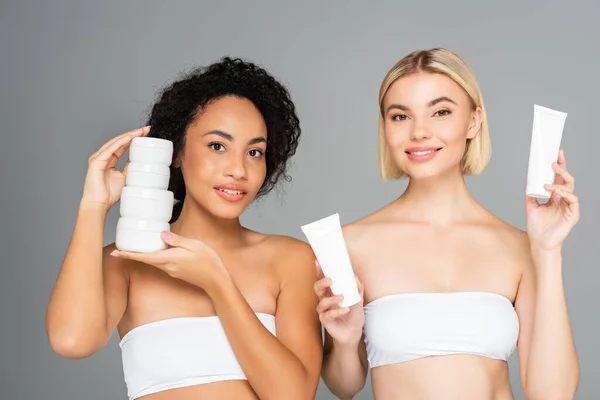 Femmes interraciales en hauts blancs tenant des bocaux et des tubes avec crème isolée sur gris — Photo de stock