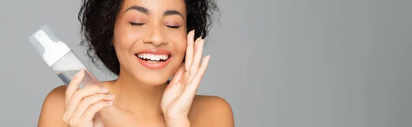 Fröhliche afrikanisch-amerikanische Frau hält Flasche Reinigungsschaum isoliert auf grau, Banner — Stockfoto