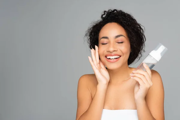 Alegre mujer afroamericana con los ojos cerrados sosteniendo botella de espuma limpiadora aislada en gris — Stock Photo