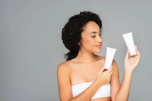 African american woman in white top holding tubes with cosmetic cream isolated on grey — Stock Photo