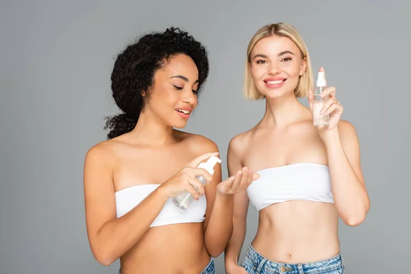 Femmes multiethniques gaies tenant des bouteilles avec mousse nettoyante et vaporisateur isolé sur gris — Photo de stock