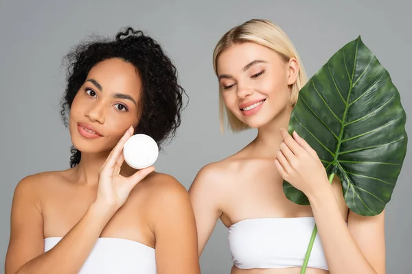 Smiling interracial women posing with cosmetic cream and tropical leaf isolated on grey — Stock Photo