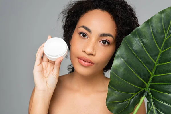 Mujer afroamericana con piel perfecta sosteniendo hoja tropical y crema cosmética aislada en gris - foto de stock