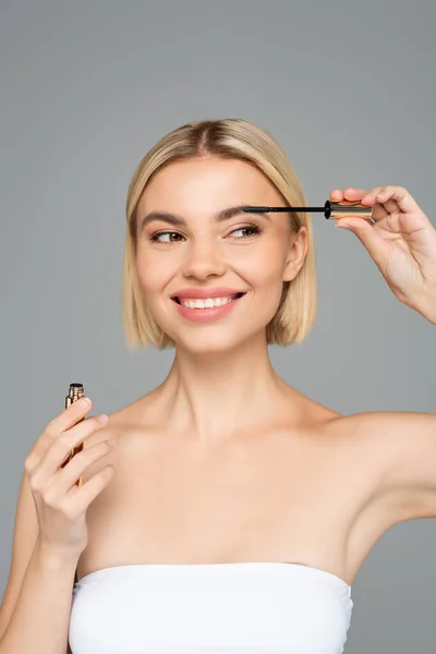 Young blonde woman holding mascara and smiling isolated on grey — Stock Photo