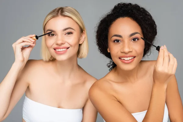 Lächelnde multiethnische Frauen, die Mascara isoliert auf grau auftragen — Stockfoto
