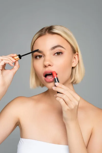 Blonde Frau trägt Lippenstift auf und hält Mascara-Applikator isoliert auf grau — Stockfoto