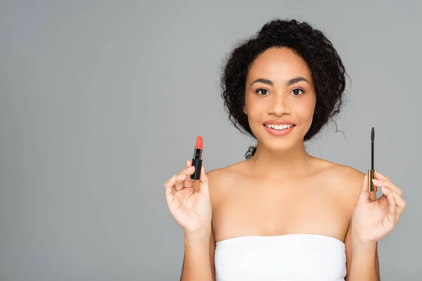 Smiling african american woman holding red lipstick and mascara isolated on grey — Stock Photo