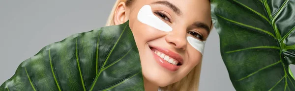 Mulher sorridente em manchas nos olhos olhando para a câmera perto de folhas tropicais isoladas em cinza, banner — Fotografia de Stock