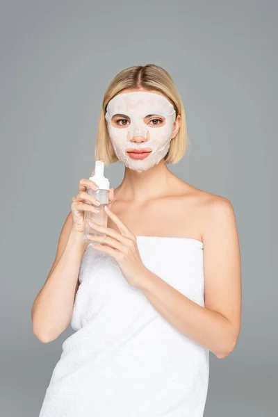 Женщина в полотенце с маской на лице, держащая очищающую пену, изолированную на сером — стоковое фото