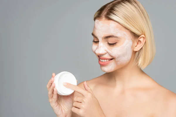 Smiling blonde woman with facial mask holding jar with cream isolated on grey — Stock Photo